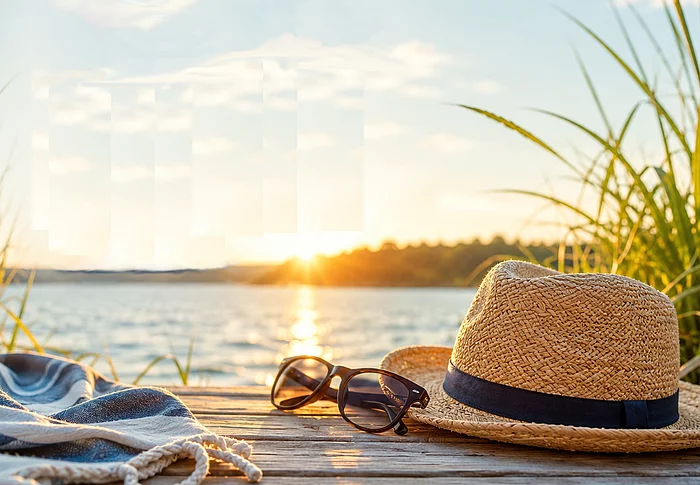 Ein Strohhut und eine Sonnenbrille auf einem Steg am See im Sonnenuntergang mit der Aufschrift „Schöne Ferien!"